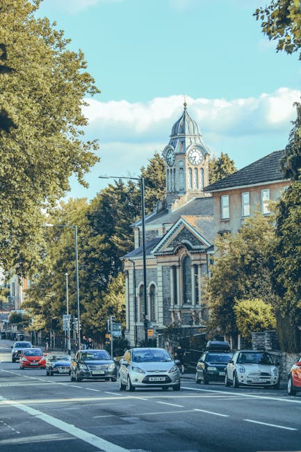 A busy street scene taken during daylight hours showing several cars parked along the curb and others driving past. To the right of the image, there is a partly visible building with a sloped roof and multiple windows, partly obscured by large, leafy green trees. In the background, a historic clock tower with a domed roof and a clock face on each side stands prominently above the surrounding structures. The sky is mostly clear with some scattered clouds, and the sunlight casts shadows on the street and buildings. The scene captures an urban environment typical of a house or apartment relocation area, where Balham Removals might assist in packing, loading, and transporting furniture and other household items through narrow streets or urban roads during house removals or moving services.