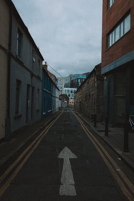 A narrow urban street during overcast weather, with a vertical lane marked by double yellow lines on each side and a white arrow painted on the asphalt indicating the direction of traffic flow. On the left, there are low-rise buildings with a mix of grey and blue facades, some with small windows and doors, and on the right, a taller modern building with large glass windows and a brick exterior. The street is used for moving furniture and packing materials during a home relocation, with no vehicles or people visible, but possibly suitable for loading or unloading during a house removal. The scene is part of the Balham High Road removals guide, illustrating the tight urban street environment encountered during narrow street moves, and may involve loading furniture into a van parked nearby, as coordinated by Balham Removals. The environment is lit by natural daylight with a cloudy sky overhead.